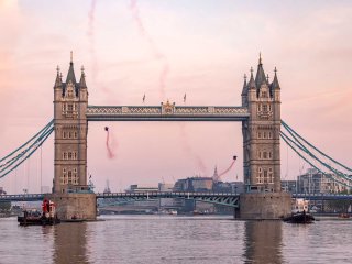 Two men in wingsuits fly through Tower Bridge, between the Walkways and the Bascules, with a pink sunrise in the background