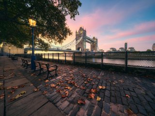 A street with autumn leaves on the ground and a street light on in the ground, with Tower Bridge in the background surrounded by pink and blue sunrise skies